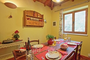 A dining table set with plates, glasses, and a bowl of fruit, with a wooden cabinet above and a window with curtains to the right. - Kitrini Luxury, Shared pool, A/C, private gardens, near beach (Ozias Gaios)