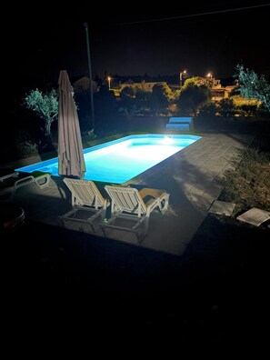 Una piscina con lettini e un ombrellone, circondata da un’area pavimentata e alberi.