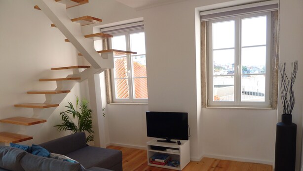 A modern living room with a grey sofa, wooden staircase, and a flat-screen TV on a white stand.