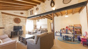A spacious living room with a stone wall, wooden beams, a television, a sofa, a coffee table, and a children's play area with toys and a bookshelf. - House (full rental) Entre Hoces for 10-12 people (Campo de San Pedro)