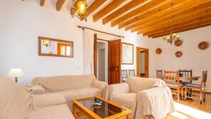 Uma sala de estar acolhedora com teto de madeira, mesa de centro de vidro, sofás bege e uma área de jantar com cadeiras e mesa de madeira.