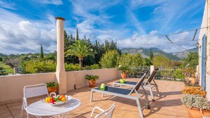 Um terraço com mesa, cadeiras, espreguiçadeira e plantas em vasos.
