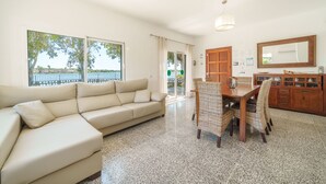 Een woonkamer met een beige bank, een eettafel met rieten stoelen, een houten kast en een grote spiegel aan de muur.