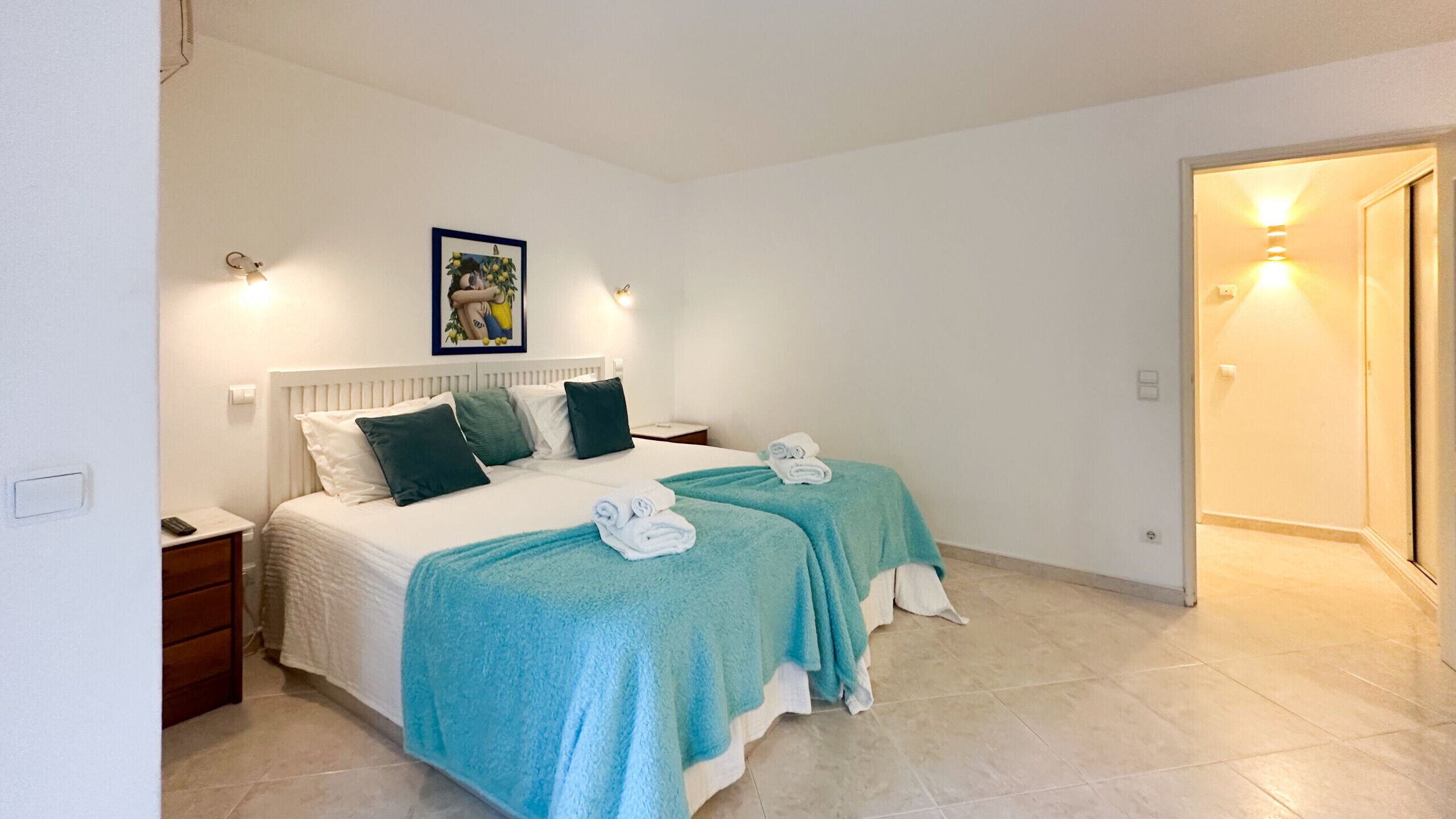 Een tweepersoonskamer met twee bedden, elk met een blauwgroene deken, witte lakens en een lijst met een foto aan de muur.