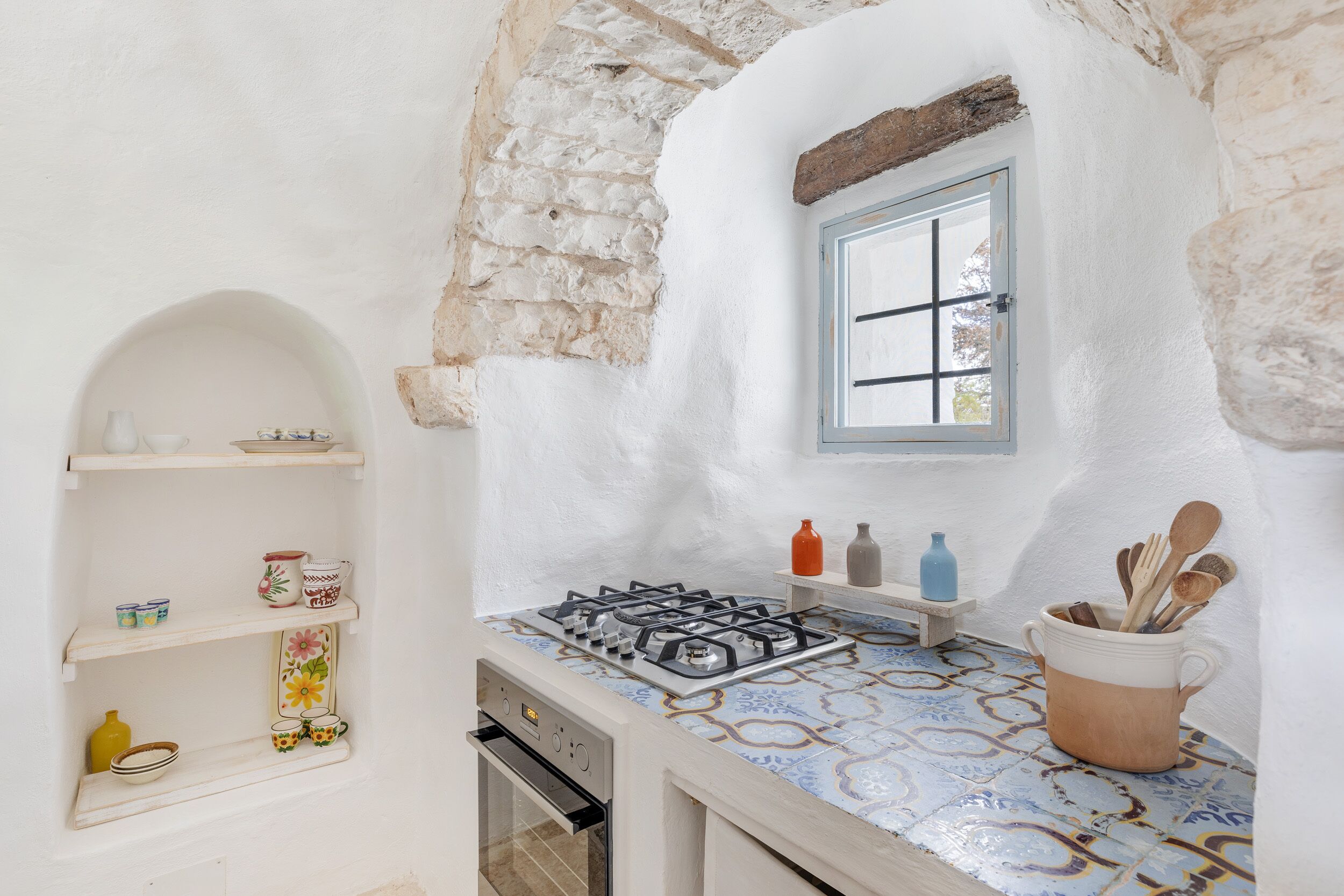 Une cuisine équipée d’une cuisinière à gaz, d’étagères remplies d’ustensiles de cuisine, d’une fenêtre et d’un mur en pierre.