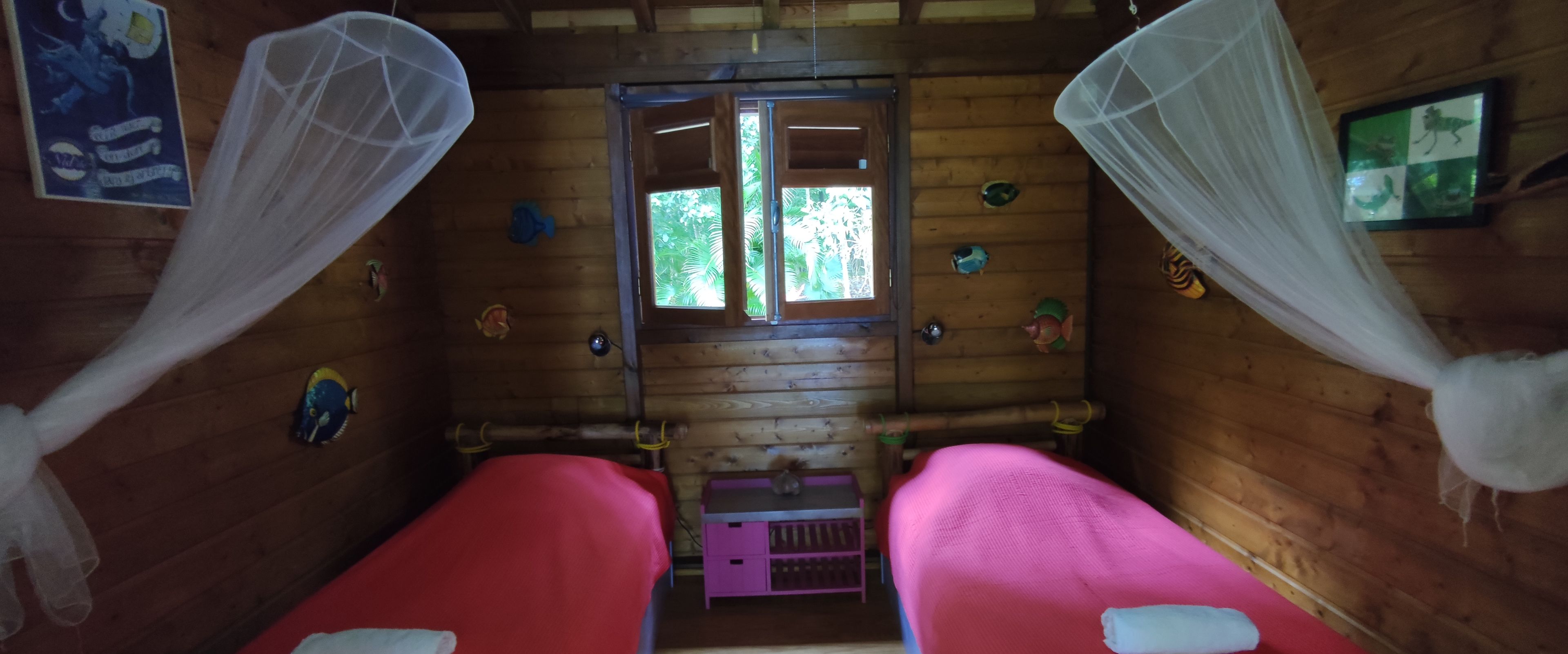 A wooden cabin room with two beds, mosquito nets, and a window with a view of greenery.