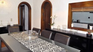 A dining area with a long table, chairs, and a decorative centerpiece. There are two arched doorways and a window with a view of the kitchen.