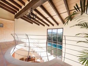 A spacious interior with a high ceiling, a staircase leading to a balcony, and a view of a swimming pool.