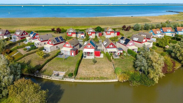 An aerial view of a residential area with houses arranged in rows, a body of water in the foreground, and wind turbines in the background.