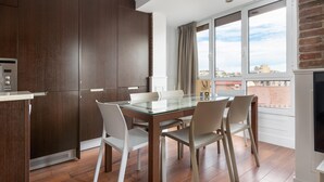 Un comedor moderno con una mesa y sillas de vidrio, una cocina con armarios de madera oscura y vistas a la ciudad a través de amplias ventanas.