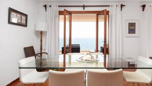 A glass-top dining table with four chairs, a lamp, and a view of the sea through a doorway.