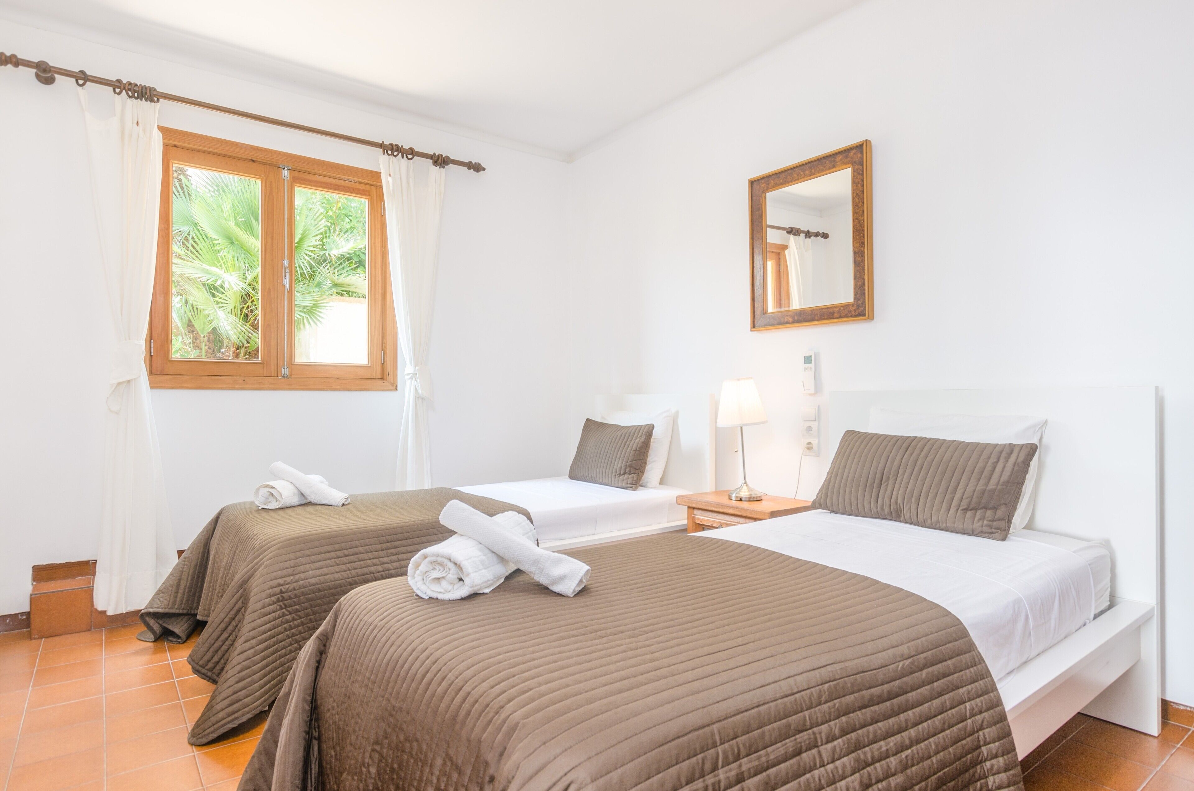 Habitación de hotel con dos camas, una ventana con cortinas, una mesita de noche con lámpara y un espejo en la pared.