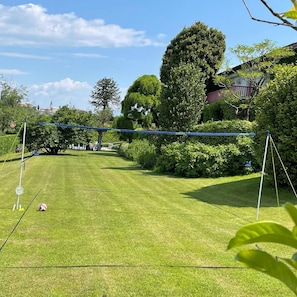 Une pelouse bien entretenue, agrémentée d’un filet de volley-ball et de poteaux de but, le tout entouré d’une végétation luxuriante et d’arbres.