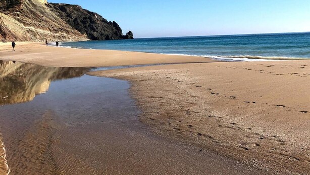 A beach with clear skies, calm waters, and a rocky cliff in the background.