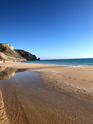 A beach with clear skies, calm waters, and a rocky cliff in the background.
