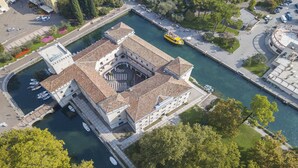 Vue aérienne d’un bâtiment historique doté d’une cour intérieure, situé à côté d’un canal, entouré de verdure et comprenant un espace de stationnement.