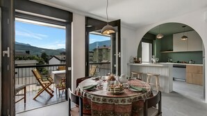 Un coin repas avec une table dressée pour quatre personnes, une cuisine avec une porte en arc de couleur verte et un balcon offrant une vue sur les montagnes.