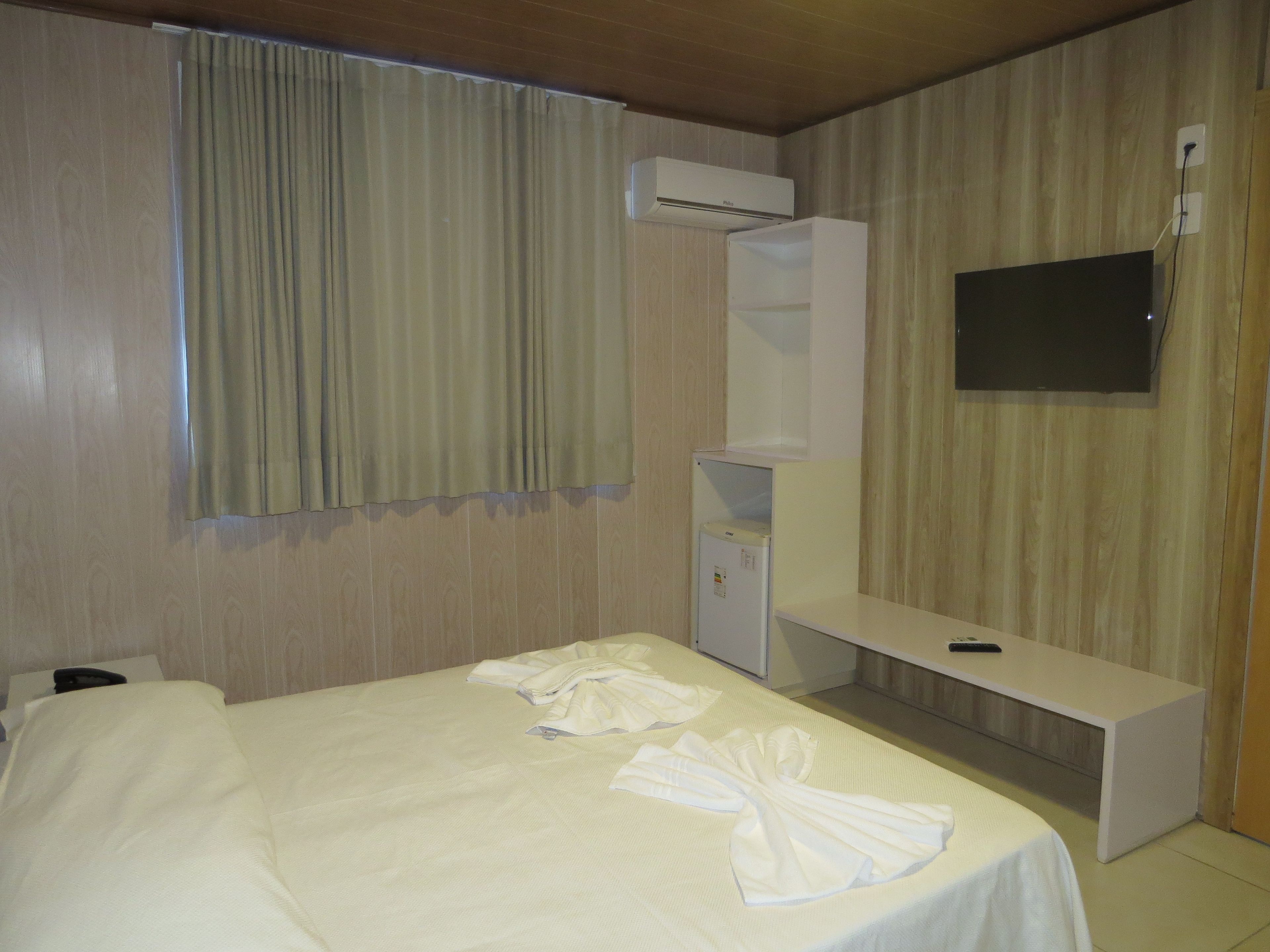 A hotel room with a bed, a desk, a flat-screen TV, and an air conditioning unit.