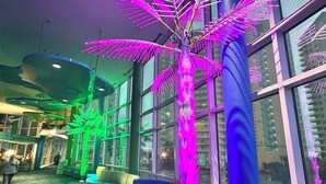Un espacio interior moderno con una gran escultura iluminada de palmera, zonas de estar y ventanas amplias que ofrecen vistas al exterior.
