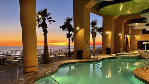 Zona de piscina con vista al atardecer sobre el océano, palmeras y sillas de descanso.