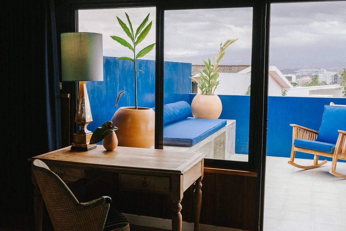 A room with a wooden desk, a chair, a sofa, a lamp, and a view of the city through large windows.