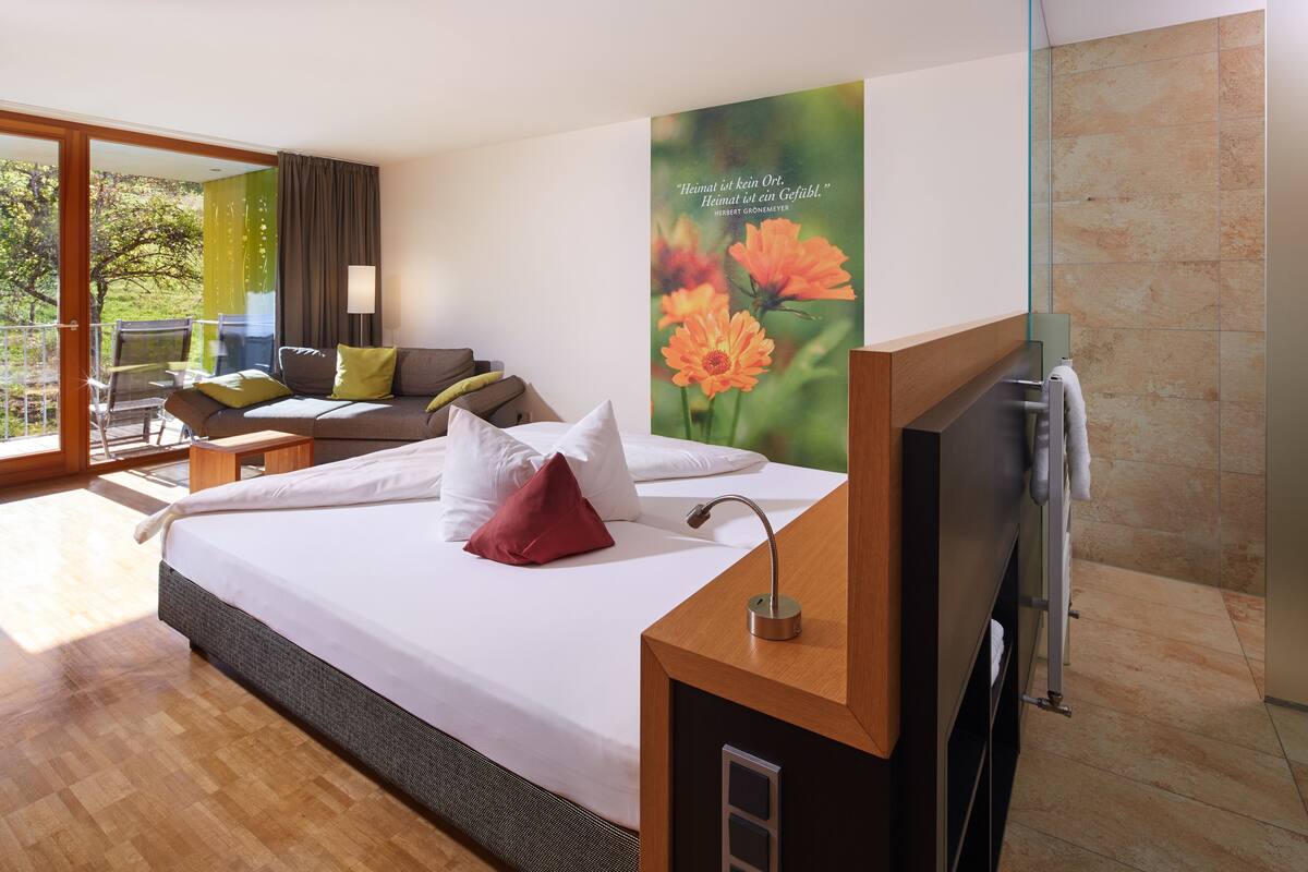 A modern hotel room with a large bed, a sofa, a glass shower enclosure, and a view of the outdoors.