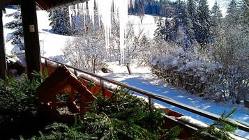 A snowy outdoor area with a wooden structure, possibly a deck or balcony, overlooking a snow-covered landscape with trees and a clear sky.