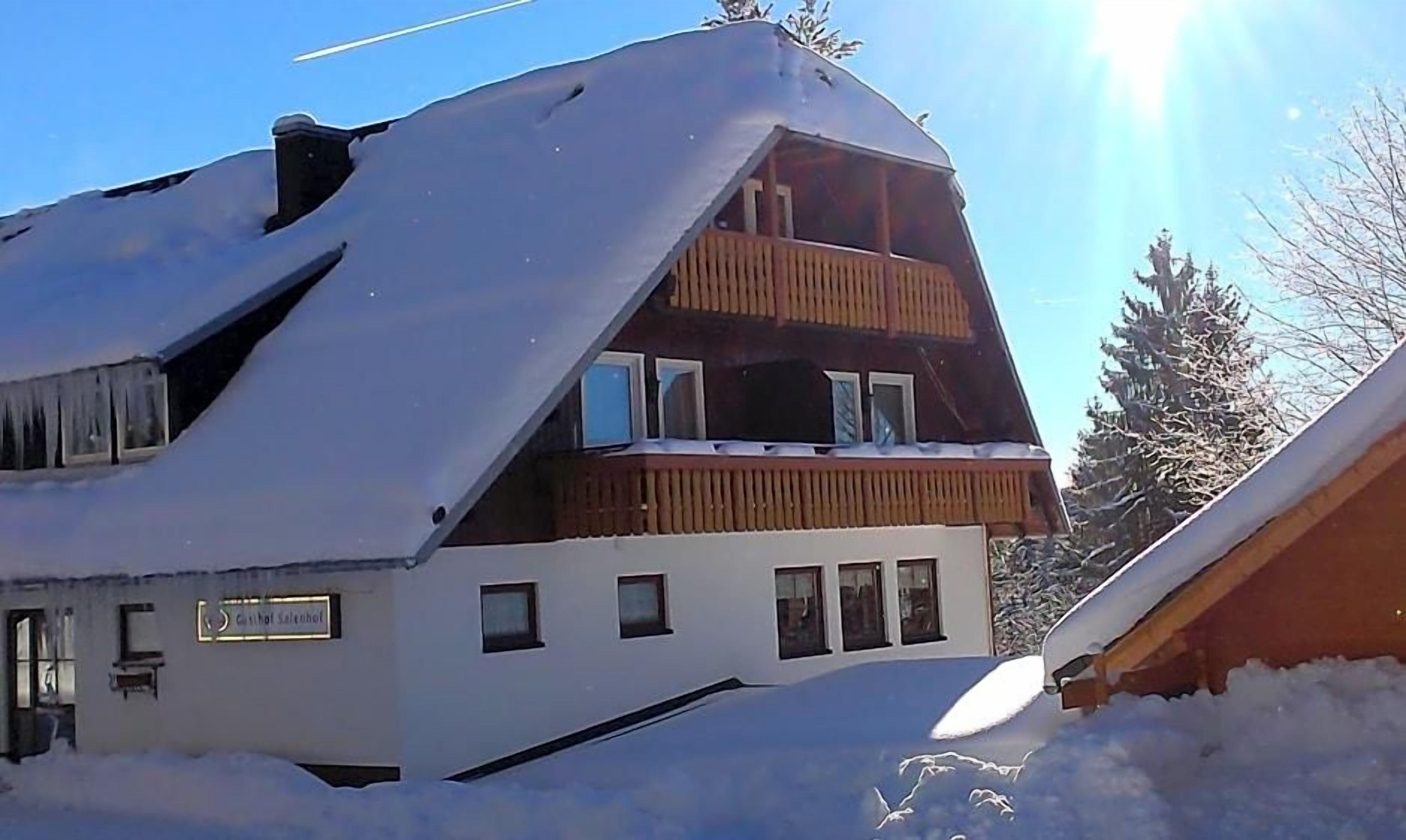 A two-story building with a sloped roof covered in snow, a wooden balcony on the upper floor, and a sign indicating it is a guesthouse.