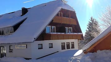 A two-story building with a sloped roof covered in snow, a wooden balcony on the upper floor, and a sign indicating it is a guesthouse.