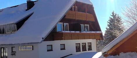 Un bâtiment à deux étages, doté d’un toit en pente recouvert de neige, d’un balcon en bois à l’étage supérieur et d’une enseigne indiquant qu’il s’agit d’une maison d’hôtes.