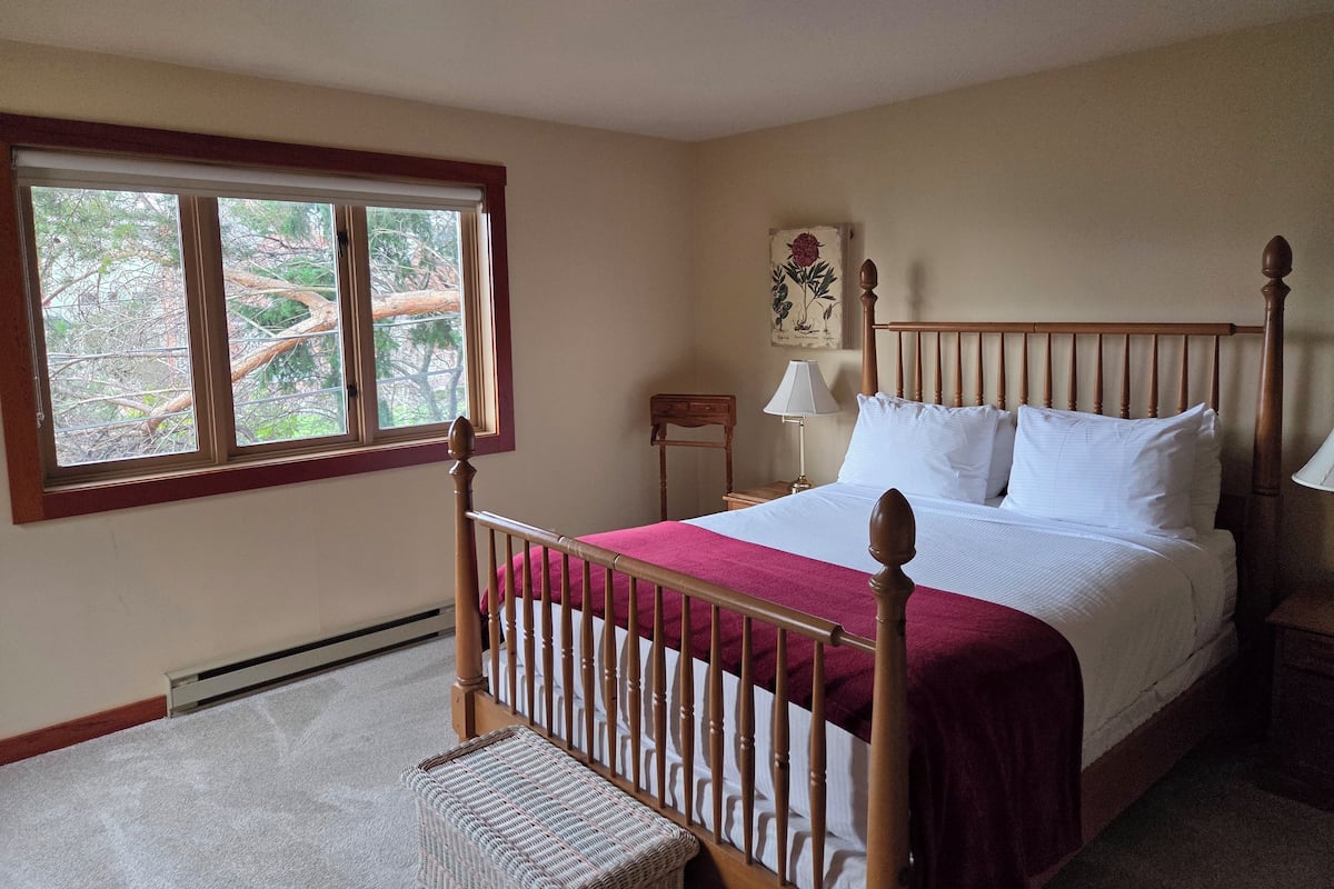 A bedroom with a wooden bed frame, white bedding, a red throw blanket, a nightstand with a lamp, and a window with a view of trees.