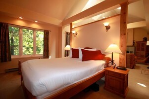 Una cama grande con ropa de cama blanca y almohadas rojas, cabecera de madera, dos lámparas de noche y vista a árboles por la ventana.