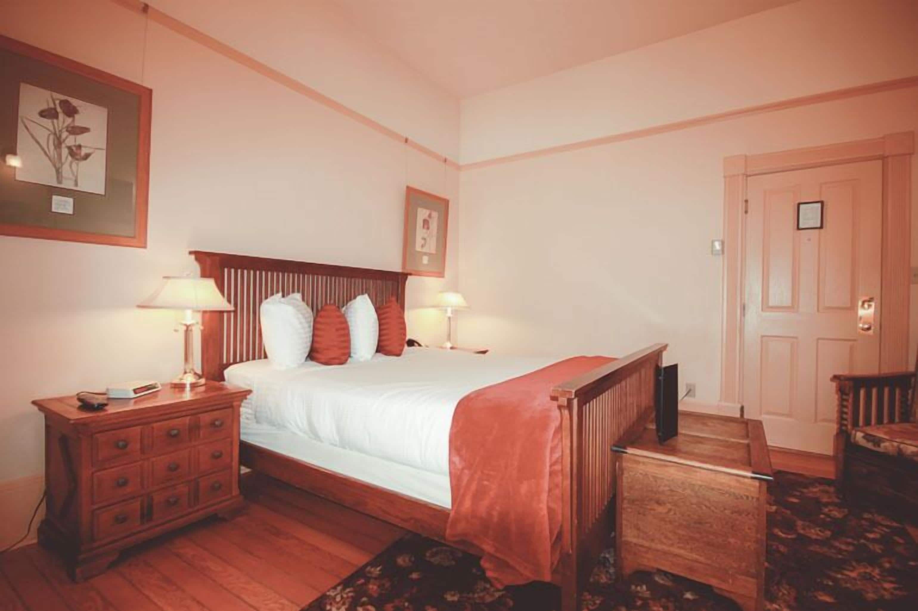 Une chambre à coucher avec un lit en bois, deux tables de chevet avec des lampes, une petite table de nuit en bois, une chaise et une porte.
