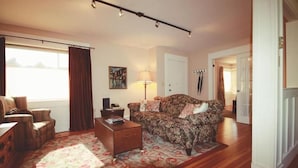 A living room with a sofa, armchair, coffee table, and a lamp. There is a window with curtains, a painting on the wall, and a door leading to another room. - The Catherine St Guesthouse (Victoria)