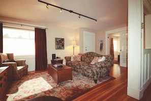 Un salón con sofá, sillón, mesa de centro y lámpara. Hay una ventana con cortinas, un cuadro en la pared y una puerta que da a otra habitación.