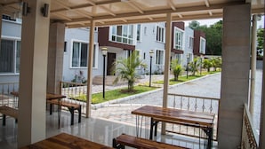 A covered outdoor seating area with wooden tables and benches, a tiled floor, and a view of residential buildings and greenery. - Troy Serviced Apartments (Lusaka)