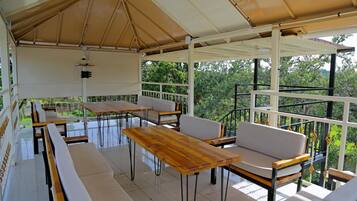 A covered outdoor seating area with tables and benches, surrounded by glass railings and greenery.