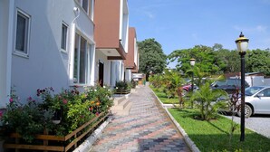 A pathway leading to a row of white buildings with brown accents, surrounded by well-maintained gardens and parked cars. - Troy Serviced Apartments (Lusaka)