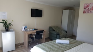 A hotel room with a bed, a desk with a chair, a television, a wardrobe, a plant, and a small refrigerator.
