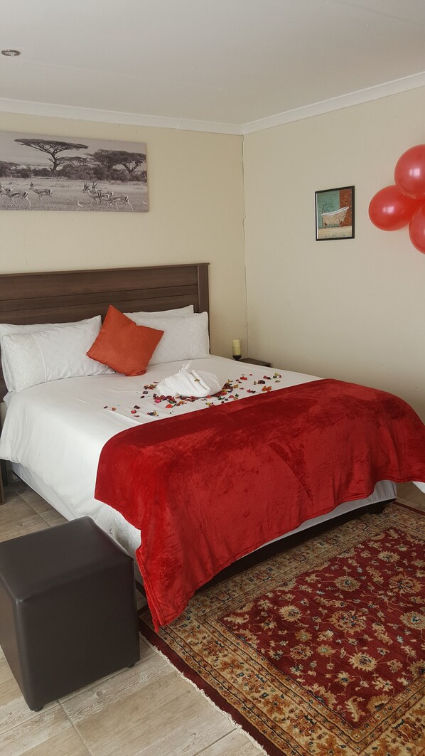A neatly made bed with a red blanket, white pillows, and a small orange pillow. A framed picture hangs on the wall above the bed, and a small table with a candle is visible in the corner.