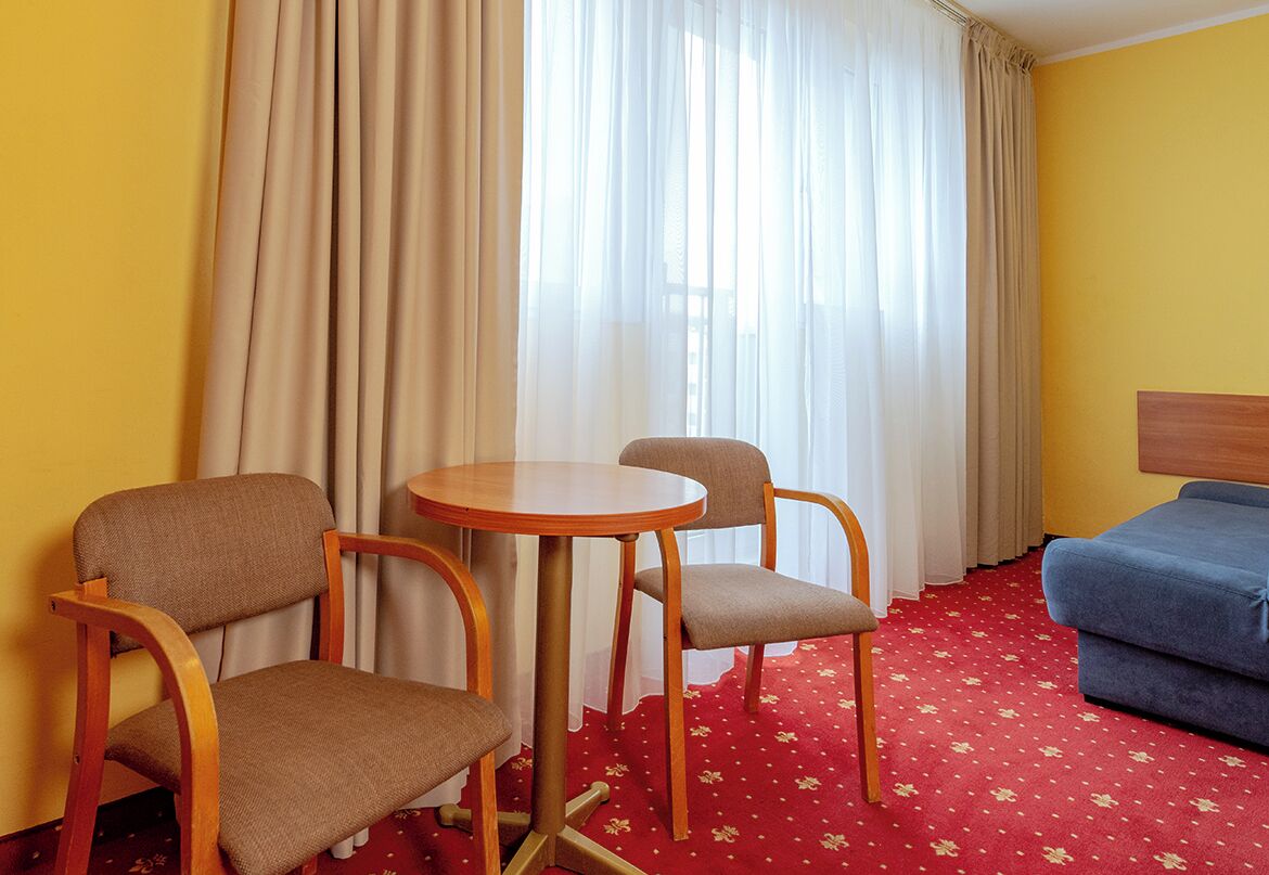 Quarto de hotel com uma mesa redonda, duas cadeiras, um sofá azul e um tapete vermelho com padrões florais.