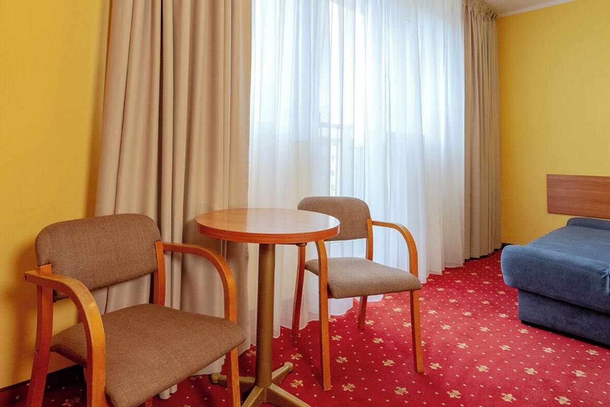 A hotel room with a round table, two chairs, a blue sofa, and a red carpet with floral patterns.