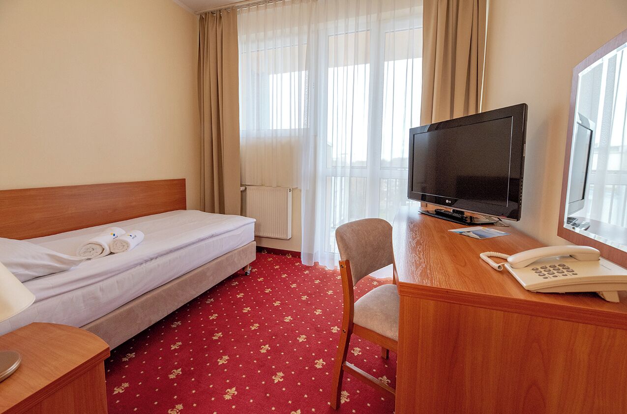 A hotel room with a bed, a desk with a TV, a chair, a phone, and a window with curtains.