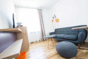 Una sala de estar moderna con un sofá, un pouf redondo, un escritorio de madera y un ventanal con cortinas.