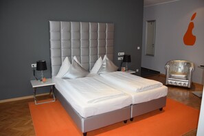 Una cama doble con cabecera capitonada en una habitación con paredes grises, una alfombra naranja y un sillón plateado.