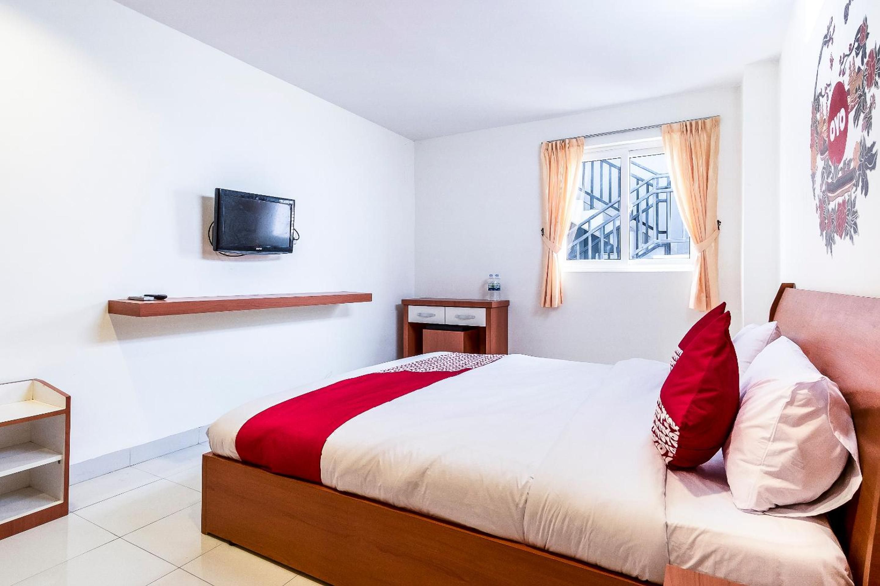 Ein Hotelzimmer mit einem Bett, einem an der Wand befestigten Fernseher, einem Fenster mit Vorhängen und einem kleinen Tisch mit einer Flasche.