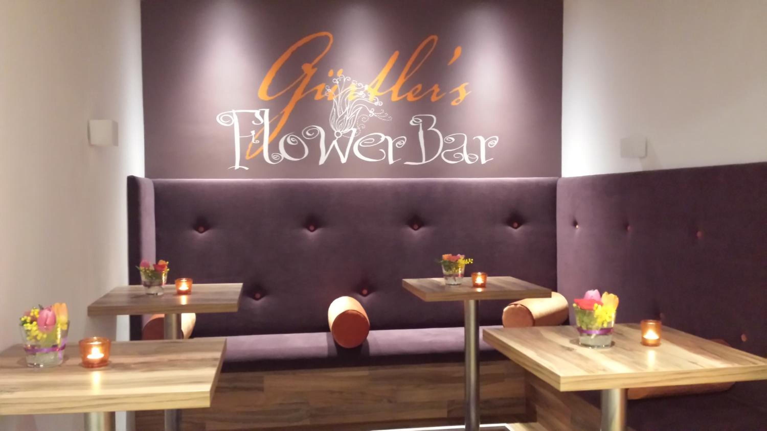 A restaurant interior with a 'Flower Bar' sign, cushioned seating, and tables with candles.