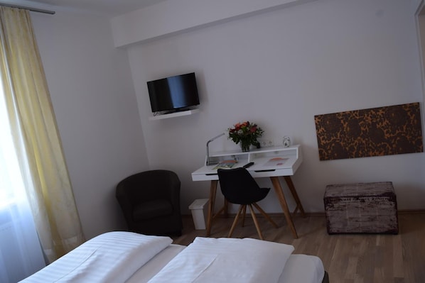 A hotel room with a bed, a desk with a chair, a television, a small table with a vase of flowers, and a painting on the wall.