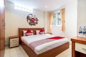 Una habitación de hotel con cama, mesita de noche, escritorio con botellas de agua, ventana con cortinas y un adorno en la pared.