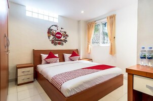 Una habitación de hotel con cama, mesita de noche, escritorio con botellas de agua, ventana con cortinas y un adorno en la pared.